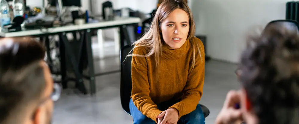 Een vrouw kijkt met een serieuze en aandachtige blik naar een collega tijdens een overleg op kantoor.